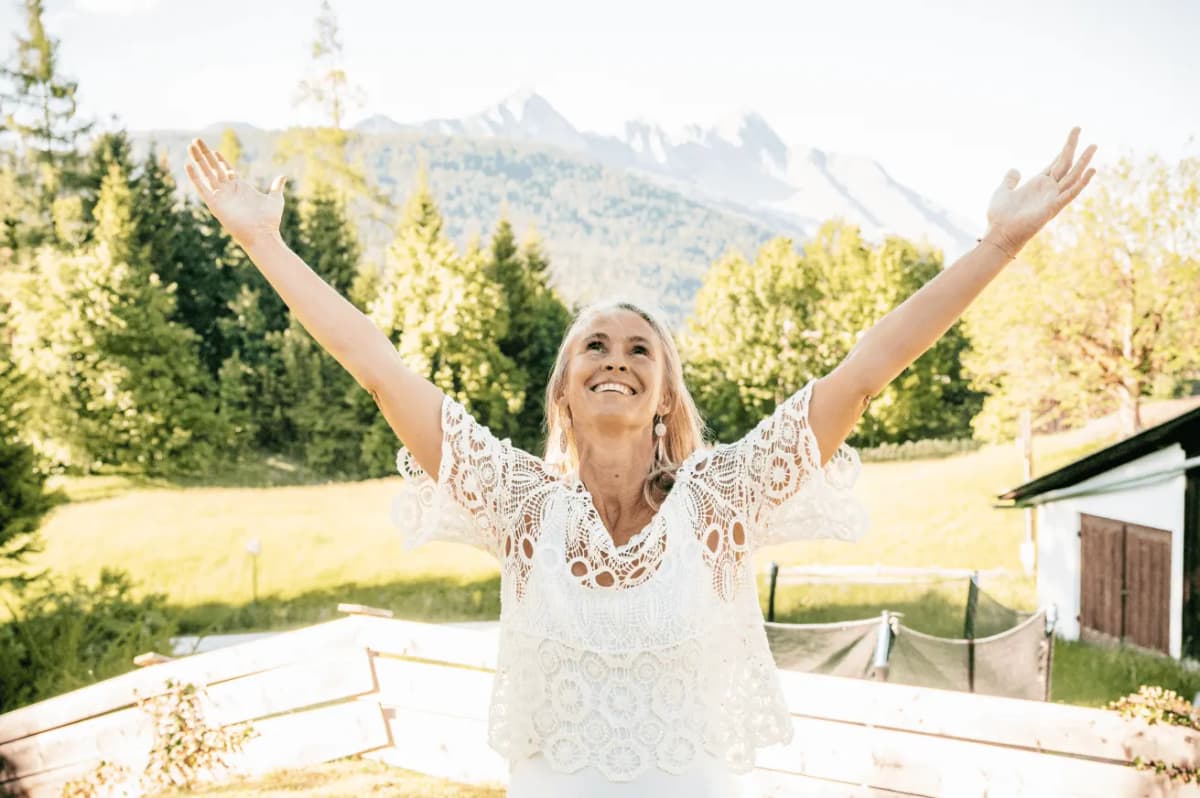 Frau in weisser Spitzenbluse streckt jubelnd beide Arme zum Himmel, im Hintergrund eine sonnenbeschienene Bergwiese und Alpengipfel