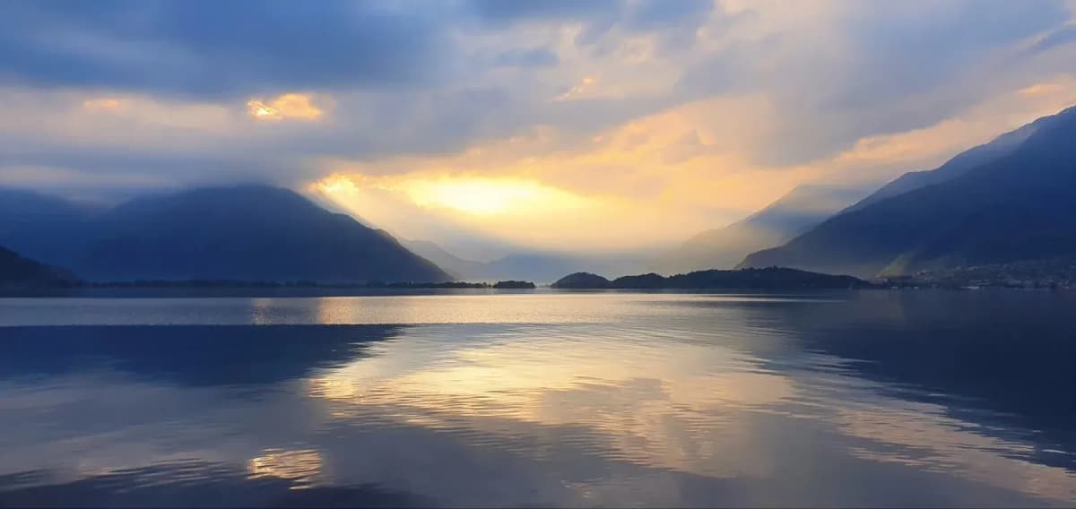 Bergsee in der Dämmerung, Sonnenstrahlen brechen durch die Wolken zwischen den Berghängen und spiegeln sich im Wasser